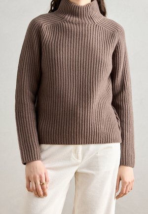 Femme portant un pull à col roulé marron côtelé à manches longues et un pantalon en velours côtelé écru, avec deux bagues en or à la main gauche.