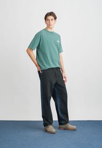 T-shirt de algodão verde com mangas curtas, combinado com jeans de denim azul escuro. Modelo usa sapatilhas de couro castanho claro. Design simples, sem padrões.