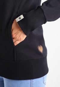 Person som bär en svart sweatshirt med handen i framfickan, visar tygetikett på ärmen och en liten lapp nära fållen, tillsammans med ljusblå jeans.