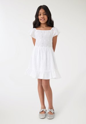 Ragazza giovane sorride indossando un abito bianco a occhielli con maniche a sbuffo e scarpe con fiocco glitter argento su sfondo bianco uniforme.