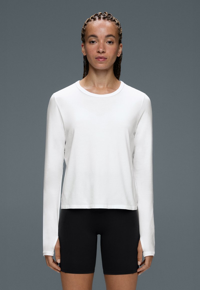 Femme aux cheveux tressés portant une chemise blanche à manches longues et un short noir, debout devant un fond gris uni.