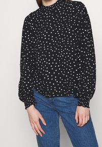 Svart blus med vita prickar, hög halsringning och rynkade puffärmar. Mjuk tyg med lös passform, parat med blå jeans.