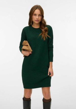 Grøn strikket sweaterkjole med rund halsudskæring, lange ærmer og en afslappet pasform, parret med en tan clutch. Tekstileret stof og knæhøje støvler.