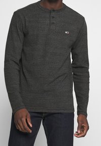 Langärmliges Henley-Shirt aus dunkelgrauem Stoff, mit Rundhalsausschnitt, dreiknöpfiger Knopfleiste und kleinem Logo auf der Brust.