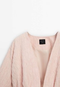 Veste texturée rose clair avec une doublure intérieure lisse et une étiquette noire indiquant Massimo Dutti Studio au niveau de l'encolure.