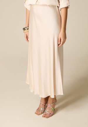 Donna che indossa una gonna midi in raso color crema, maniche arrotolate, sandali con tacco multicolore con cinturini e due braccialetti spessi su un polso.