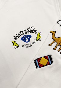 Witte katoenen t-shirt met kleurrijke borduurwerk: een blauwe aap, cactussen, een kameel en een gestreepte tapijt, met de tekst "BLCK LVNS".