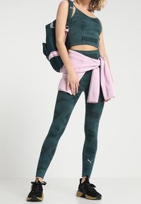 Kobieta w ciemnozielonej koszulce sportowej i legginsach w kamuflażu marki Puma, z różowym swetrem przewiązanym w talii i pasującym zielonym plecakiem.