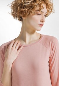 Jasnoróżowy, długi rękaw top z miękkiej, teksturowanej tkaniny z okrągłym dekoltem i rękawami raglanowymi. Modelka delikatnie dotyka obojczyka.