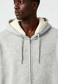 Felpa grigia con zip, dotata di un colletto foderato in pile di colore crema e cordini con punte in metallo; texture morbida e design casual.