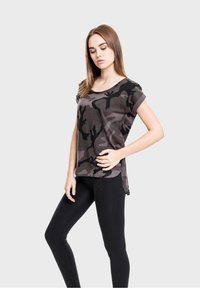 Young woman with straight brown hair wears a gray and black camouflage T-shirt and black leggings, posing against a plain light background.
