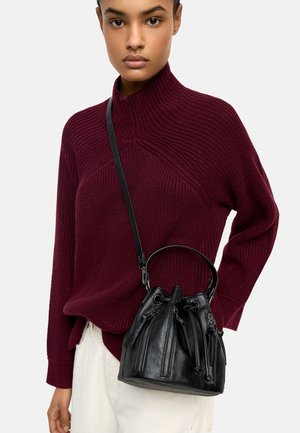 Osoba ubrana w bordowy sweter dzianinowy i kremowe spodnie, niosąca czarną skórzaną torbę typu bucket z zamkami błyskawicznymi i ściągaczem.