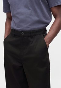 Homme portant une chemise à manches courtes gris foncé et un pantalon noir, les mains dans les poches, sur un fond clair uni.