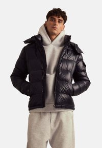 Doudoune marine avec une finition brillante, fermeture à glissière et capuche, portée sur un sweat à capuche gris, associée à un pantalon de survêtement gris clair.