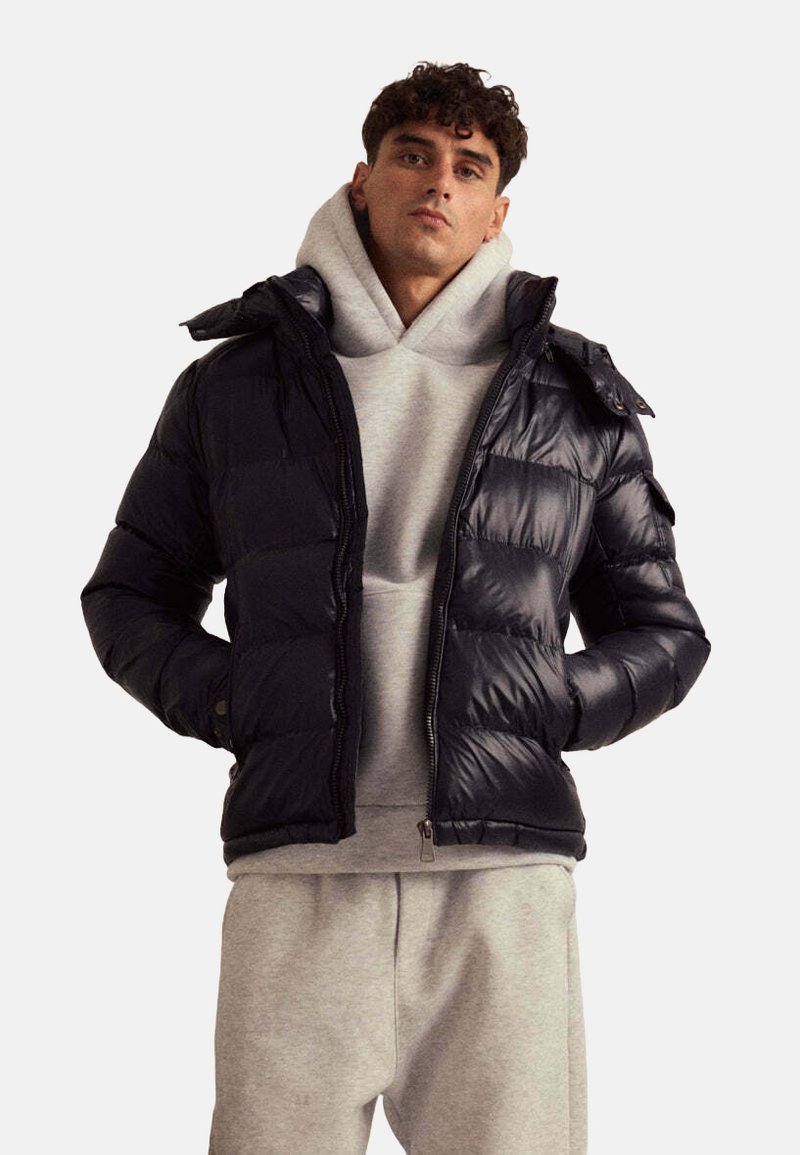 Doudoune marine avec une finition brillante, fermeture à glissière et capuche, portée sur un sweat à capuche gris, associée à un pantalon de survêtement gris clair.