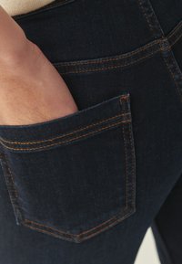 Sötét farmernadrág narancssárga varrással. Közeli kép egy kézről, amely részben a hátsó zsebben van, kiemelve a zseb méretét és formáját.