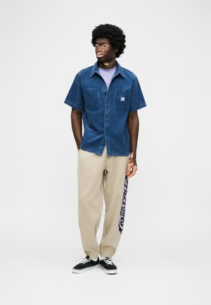 Noor vitiligoga noormees, kes kannab sinist lühikeste varrukatega särk, beeže kõrvalmustriga pükse ja musta spordijalatsit, seisab valge tausta ees.