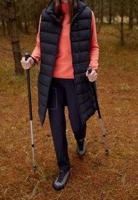 Svart vadderad väst över en korallfärgad zip-up-tröja, kombinerat med mörka byxor och grå vandringsskor. Två vandringsstavar i handen, omgiven av tallbarr.