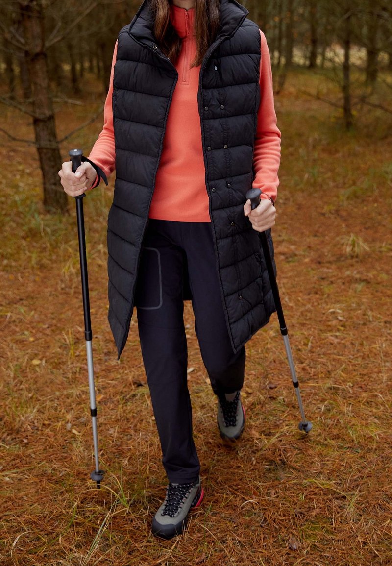 Svart vadderad väst över en korallfärgad zip-up-tröja, kombinerat med mörka byxor och grå vandringsskor. Två vandringsstavar i handen, omgiven av tallbarr.