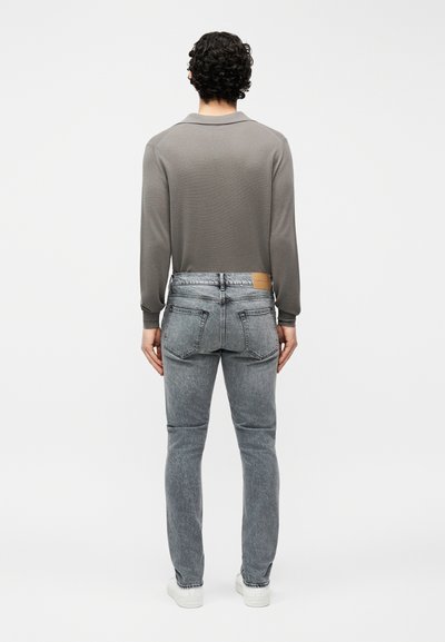 Άνδρας με σγουρά μαλλιά που φορά γκρι μπλούζα με μακριά μανίκια, ξεθωριασμένο γκρι τζιν και λευκά αθλητικά παπούτσια, στέκεται γυρισμένος με την πλάτη προς το μέρος μας σε απλό λευκό φόντο.