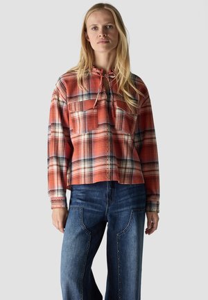 Woman wearing an orange plaid long-sleeve shirt with front pockets and blue jeans with seam details, standing against a plain background.