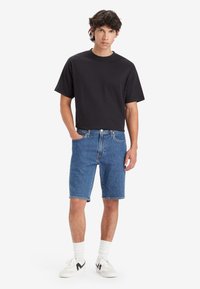 Tricou negru cu mânecă scurtă, cu logo brodat, asortat cu pantaloni scurți din denim albastru. Modelul poartă adidași albi și șosete de lungime medie.