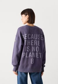 Purpurinė džemperis su pertekliniais siluetais, su dideliu reljefiniu tekstu ant nugaros: "DAILIAI, KAD NĖRA PLANETOS B." Ribbami rankogaliai ir apykaklė.