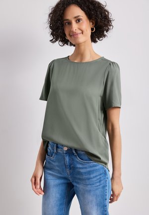 Mujer con cabello rizado y oscuro lleva una blusa verde de manga corta y vaqueros azules, de pie frente a un fondo claro y liso, sonriendo ligeramente.