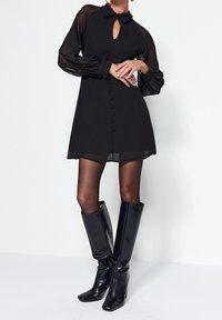Schwarzes Kleid mit transparenten langen Ärmeln, einem Kragen und einem Schlüsselspalt-Ausschnitt. Verfügt über eine Reihe von Knöpfen auf der Vorderseite. Schwarze kniehohe Stiefel.