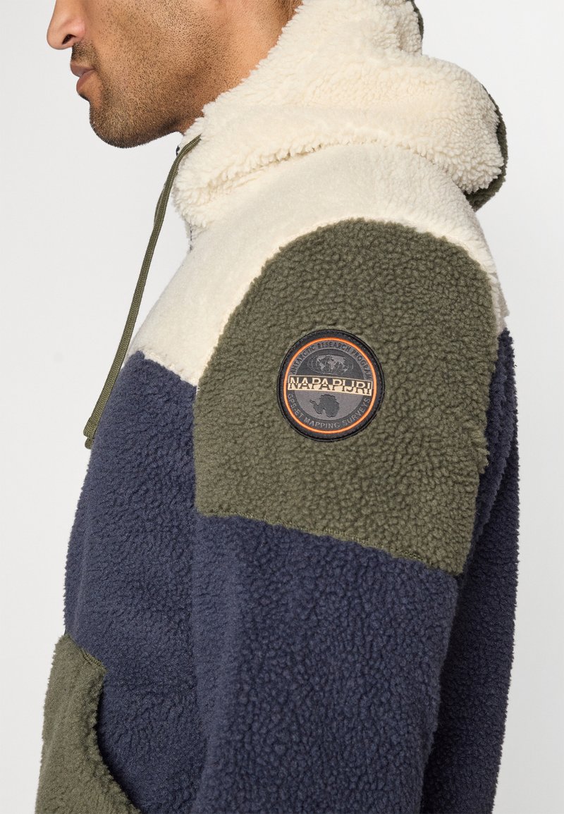 Mężczyzna ubrany w granatowy, oliwkowy i kremowy polarowy hoodie z okrągłym patch'em logo Napapijri na rękawie.