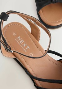 Črne zamsheve sandale s rjavim vložkom, ki imajo križaste naramnice in šivane detajle. Na vložku je logo "NEXT Forever Comfort".