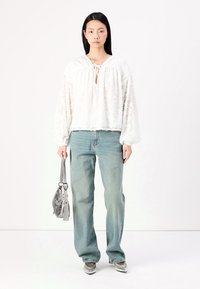 Blusa branca texturada com um laço no pescoço, combinada com jeans relaxados azul claro. Bolsa prateada e sapatos prateados de bico, de pé contra um fundo branco.