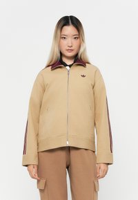 Giacca beige con colletto bordeaux, chiusura con zip e tre strisce bordeaux lungo le maniche. Presenta un piccolo logo sul petto.