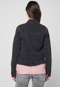 Black denim jacket with a fitted design, button detail on the sleeve, and a slightly curved hem, layered over a light pink shirt.