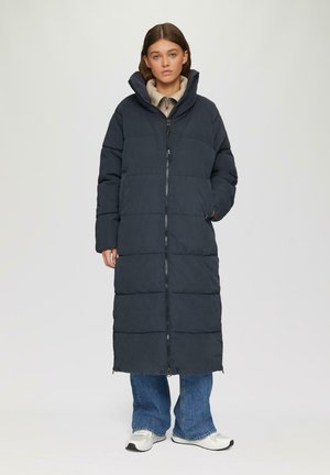 Manteau rembourré long bleu marine avec un col haut, fermeture zippée à l'avant et couture horizontale. Associé à un jean bleu et à des chaussures de sport.