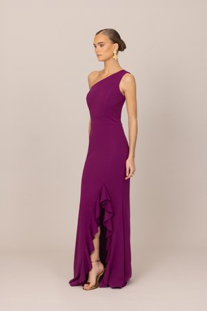 Kobieta w fioletowej sukni na jedno ramię, bez rękawów, sięgającej do ziemi, z falbankowym rozcięciem z przodu, stojąca bokiem, w złotych sandałach na wysokim obcasie.