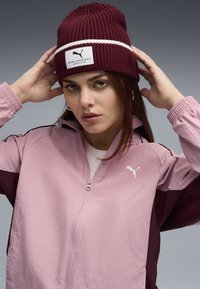Giacca zip-up di colore rosa chiaro con accenti marrone scuro e logo Puma. Berretto lavorato a maglia bordeaux con striscia bianca e etichetta Puma Athletics.