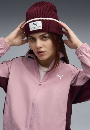 Lichtroze zip-up jack met donker maroon accenten en Puma-logo. Bordeaurood gebreide beanie met witte streep en Puma Athletics-label.