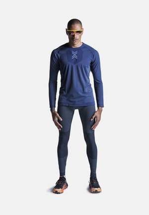 Atletische man staat, draagt een donkerblauw shirt met lange mouwen, donkere compressiebroek, zwarte en oranje sneakers en gele sportzonnebrillen.