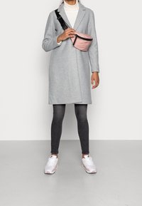 Grauer Wollmantel mit einem Schalkragen, getragen über schwarzen engen Jeans und weißen Sneakers, ausgestattet mit einer pinken Ta Tasche mit schwarzem Reißverschluss.