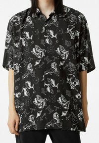 Camisa negra de manga corta con botones, con símbolos astrológicos blancos y gráficos de animales, confeccionada en una tela suave y con un corte holgado.