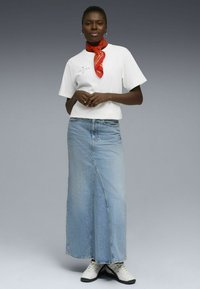 T-shirt blanc à manches courtes avec un graphisme, jupe longue en jean bleu clair avec une couture centrale, et une écharpe rouge nouée autour du cou. Chaussures blanches.