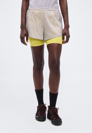 Personne portant un short beige par-dessus un cuissard jaune, des chaussettes noires et des chaussures de sport noires avec des lacets rouges, debout sur un fond blanc.