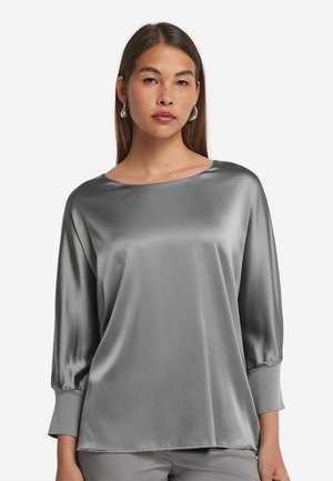 Blouse en satin gris avec des manches trois-quarts, un col rond et une texture lisse. Elle présente une coupe décontractée et une tombée douce.