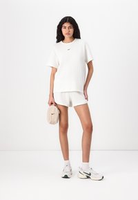 Tricou alb cu maneci scurte și textura striată, având logo-ul negru Nike, asortat cu pantaloni scurți albi. Modelul ține în mână o geantă mică crem cu logo. Poartă adidași albi.