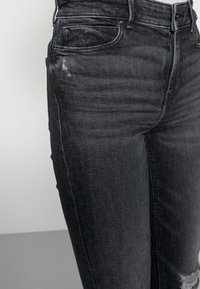 Primer plano de unos vaqueros negros desgastados con cremallera frontal y trabillas, mostrando un ligero desgaste en la parte superior del muslo y un desgarro cerca de la rodilla.