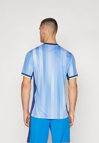 Sports jersey i lyseblå med lodrette striber, marineblå sidepaneler og korte ærmer med gule detaljer ved manchetterne.