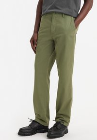 Olivenfärgade bomullsbyxor med rak passform, med en framficka, bältesöglor och bärs med svarta skinnskor.