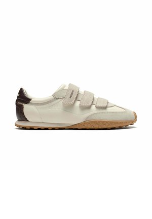 Zapatilla blanca de perfil bajo con correas de ante beige, parche marrón en el talón y suela de goma texturizada vista de lado.