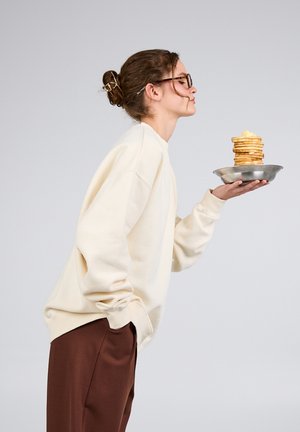 Junge Frau mit Brille und Dutt hält einen Teller mit einem hohen Stapel Pfannkuchen, die mit Butter belegt sind. Sie trägt ein cremefarbenes Sweatshirt und braune Hosen.